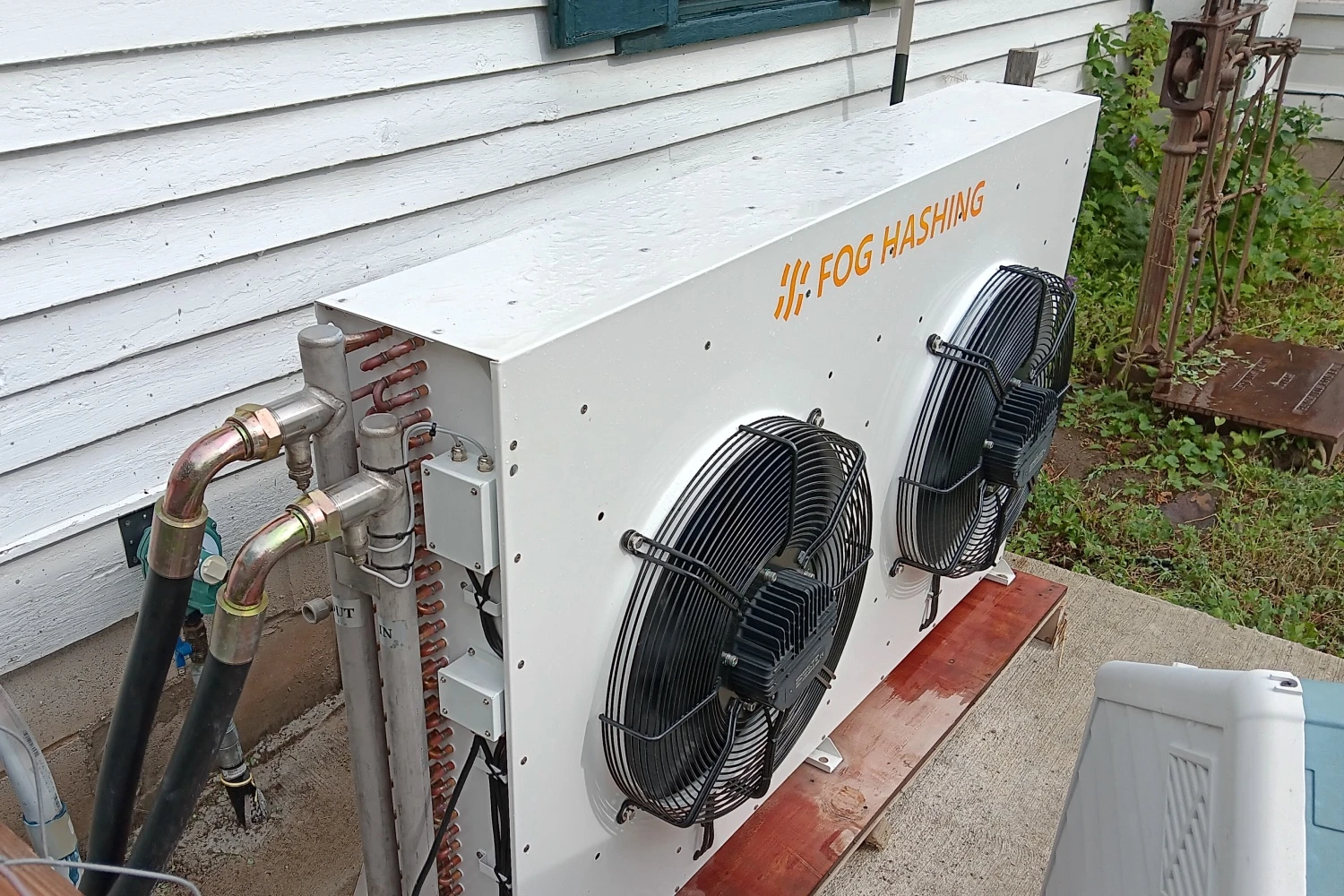 A Fog Hashing dry cooler on a pad against a clapboard house wall, copper flow and return lines below it