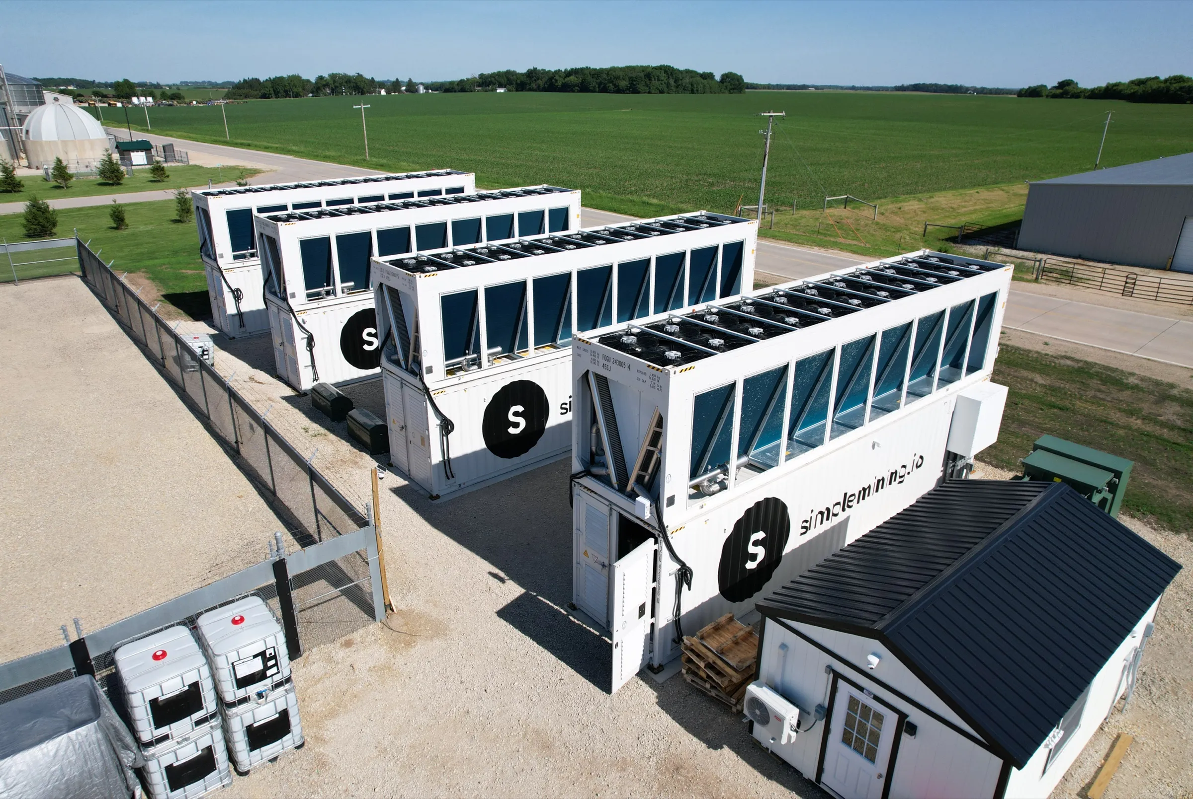 Aerial of the Simple Mining site: four Fog Hashing hydro-cooling containers on a fenced pad beside farmland