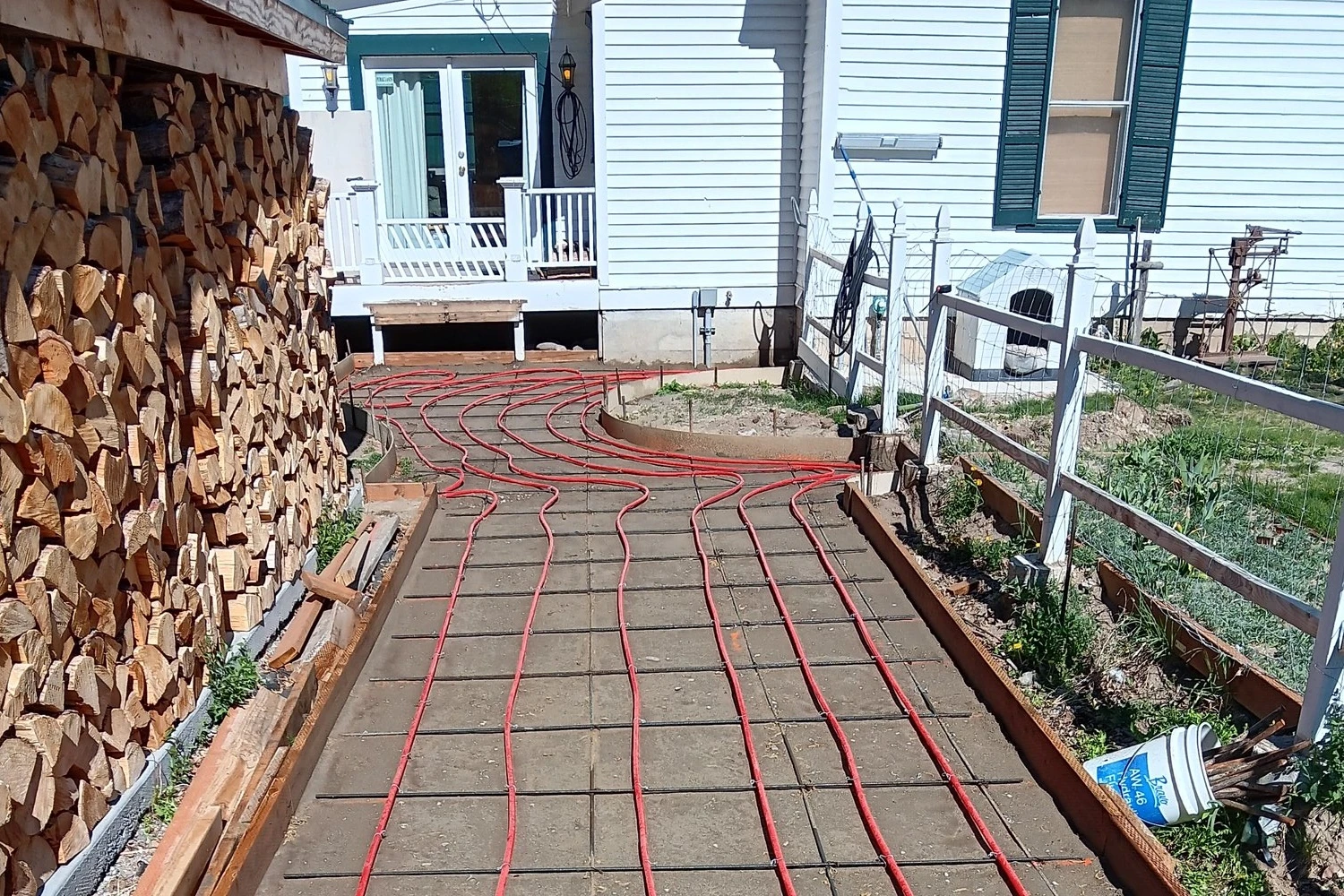 Red hydronic tubing tied to the rebar of a walkway slab before the pour, house and woodpile behind
