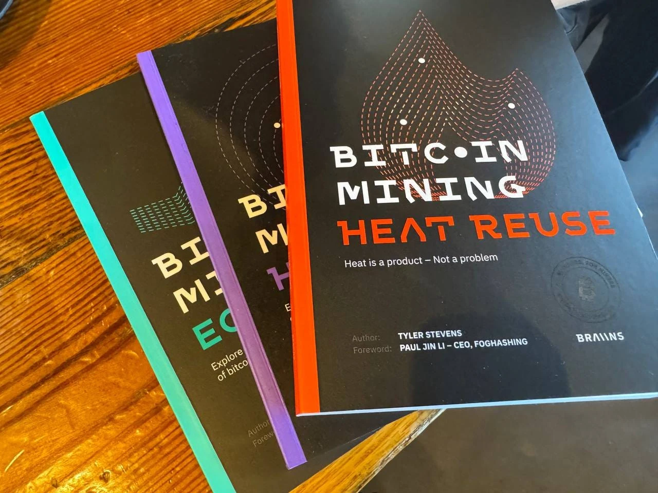 Three Braiins paperbacks on a wooden table, Bitcoin Mining Heat Reuse on top, its cover crediting Tyler Stevens as author and Paul Jin Li for the foreword