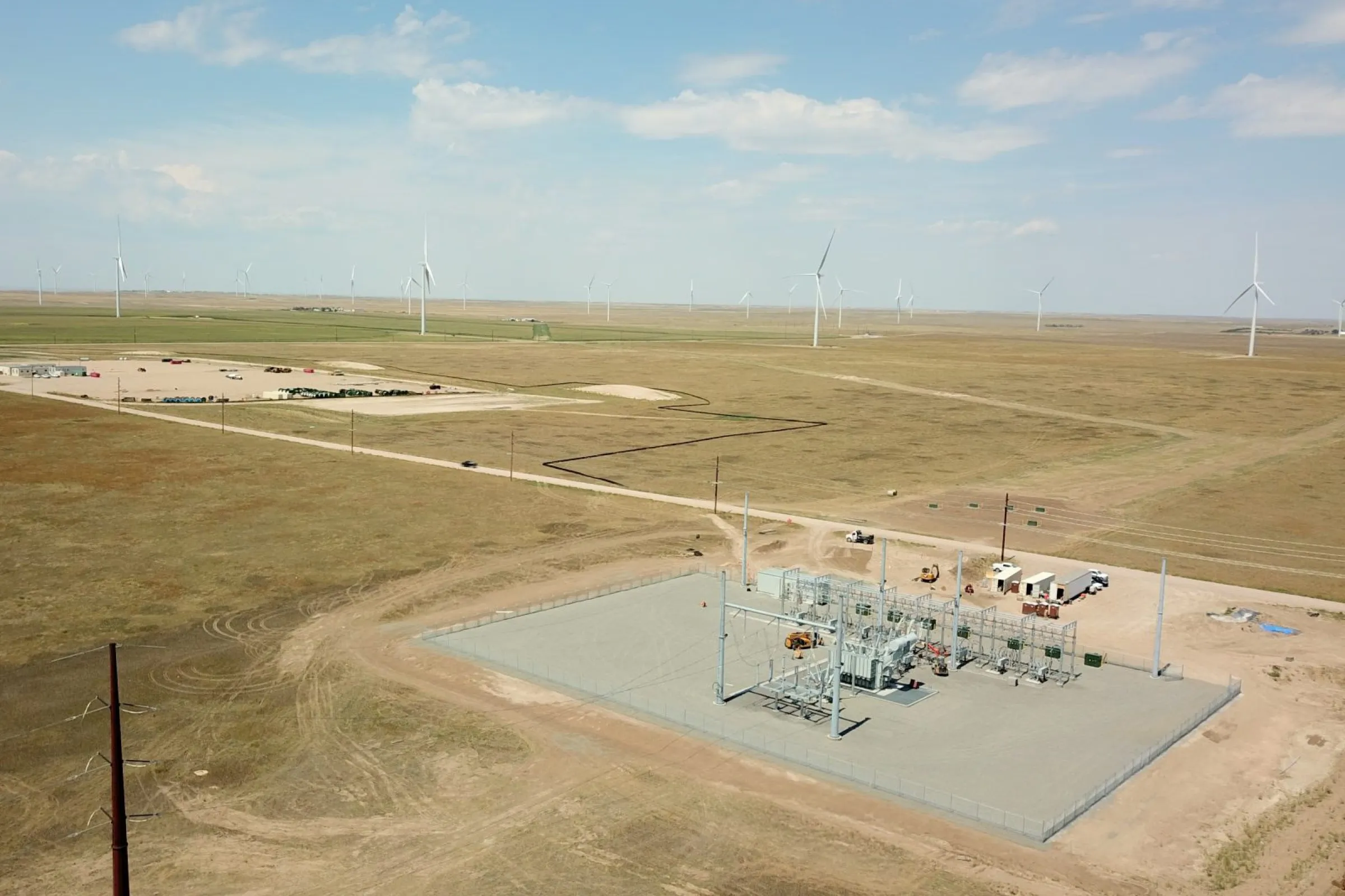 A substation on the plains seen from the air, a wind farm running to the horizon behind it
