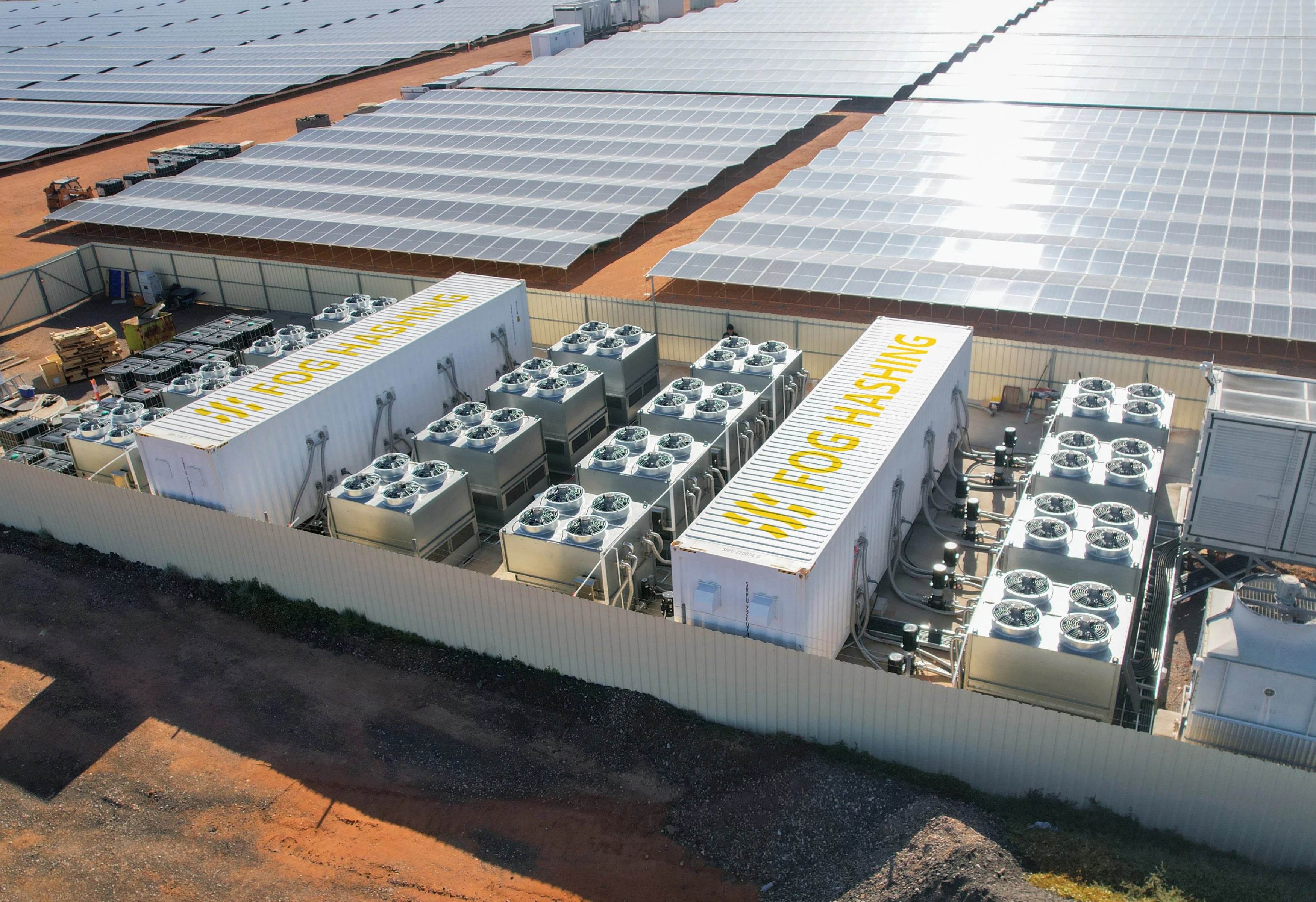 Two Fog Hashing containers and their dry cooler banks inside a fenced compound, a utility-scale solar array filling the ground behind, seen from the air